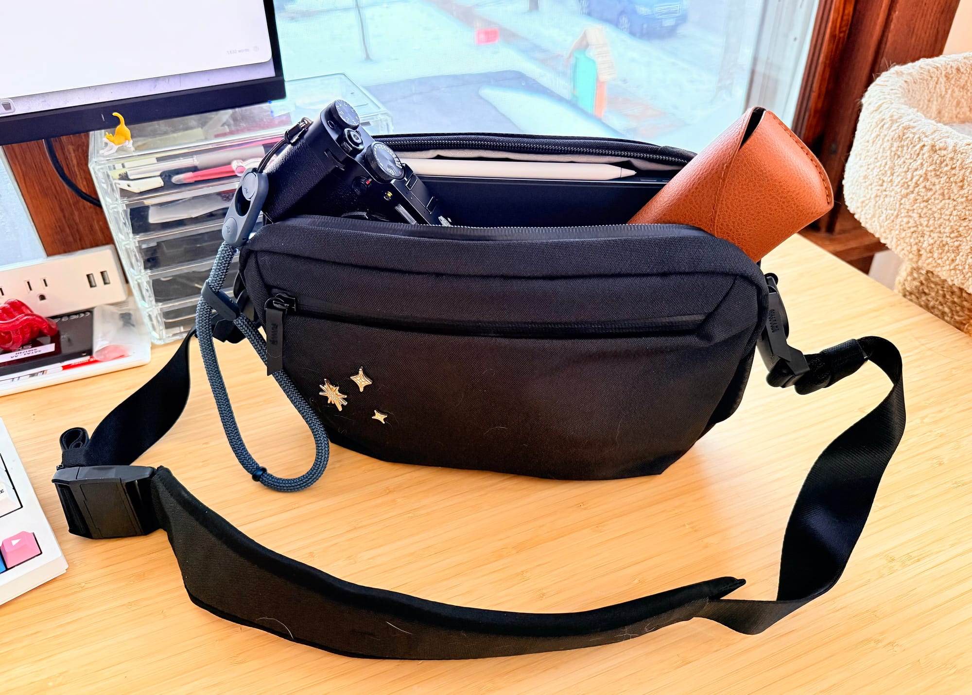 A black sling bag with a single strap sits on a wood desk. It's unzipped at the top and peaking out are sunglasses, a digital camera, and an iPad. There are three star pins on the front of the bag