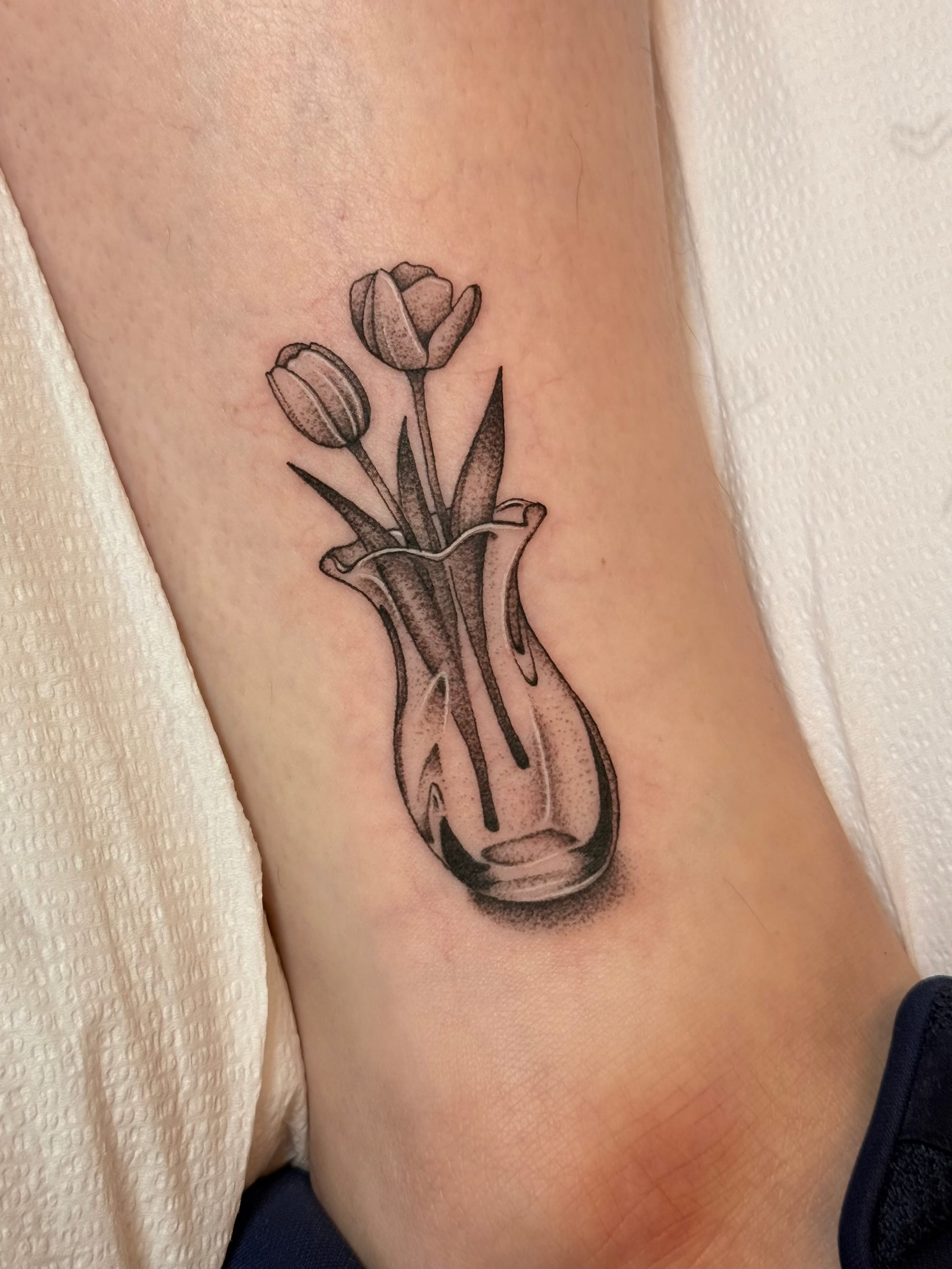 A photograph of a fresh tattoo on a woman's ankle. The tattoo is black and gray with some white highlights and is of two tulips in a vase
