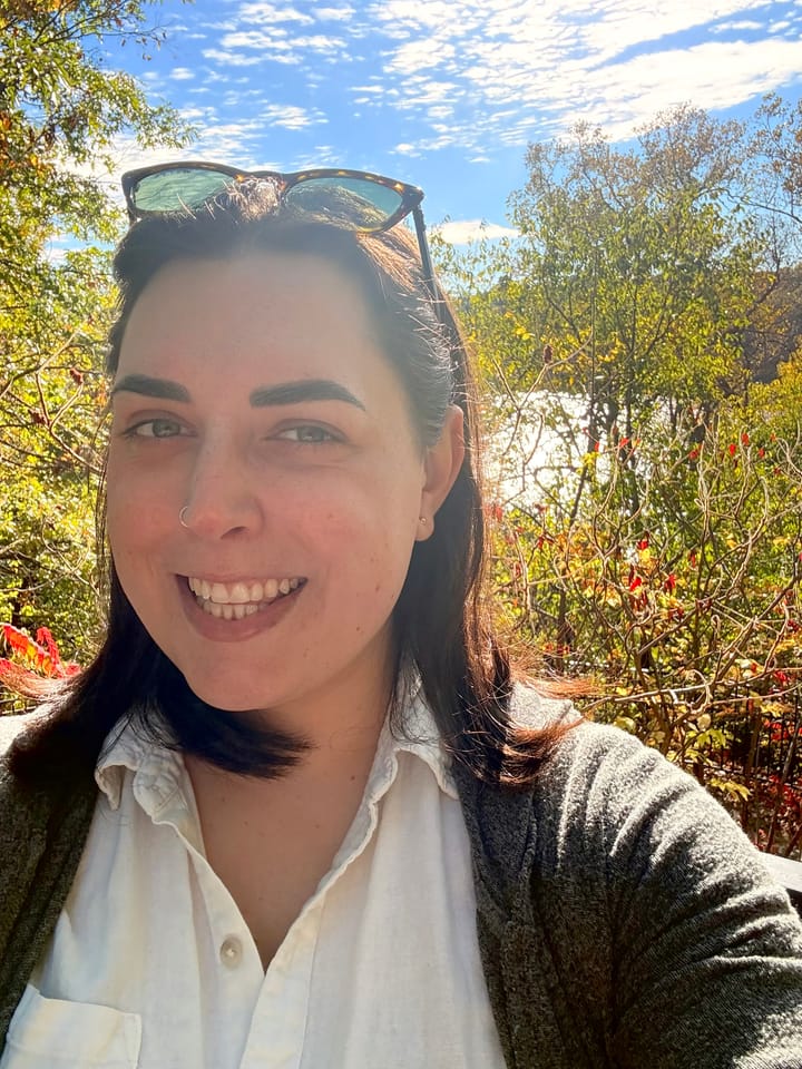 A white woman with shoulder-length brown hair smiles at the camera, she's outside with a background of trees, sky, and a peek of water