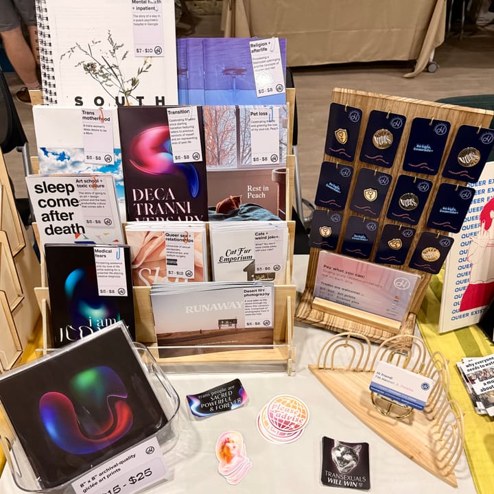 Table setup showing a display full of zines, enamel pins hanging, a payment sign, a clear box with art prints, and different sticker designs next to business cards